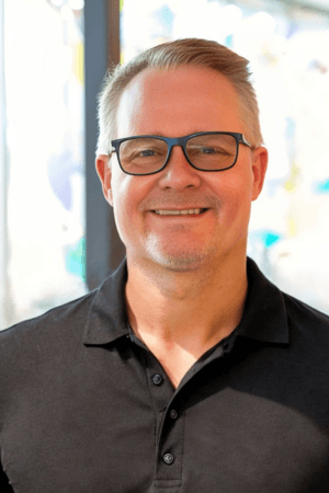 A smiling man with short hair and glasses, wearing a black polo shirt, stands confidently in front of a brightly lit, abstract background, embodying the energy of a Career Transition Coach.