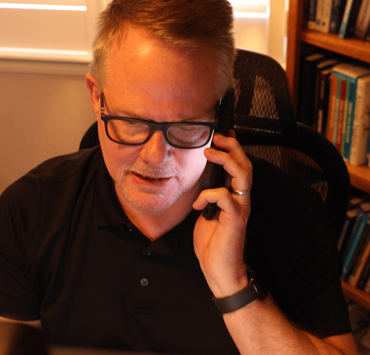 A person wearing glasses and a black shirt is sitting in a warmly lit room, reminiscent of a home office, talking on the phone. Behind them, a bookshelf filled with books hints at their expertise, possibly offering career coaching and resume services to clients seeking professional guidance.