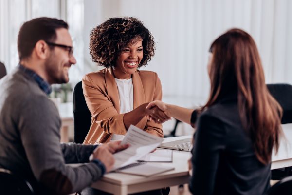 Three people are sitting at a table in an office setting. A woman in a brown blazer, perhaps a Certified Career Coach, smiles while shaking hands with another woman across from her. A man in glasses sits beside them holding documents from professional resume writing services, smiling.