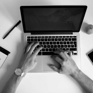 A person typing on a laptop at a desk with a smartphone, pen, notebook, and an apple nearby. The image is in black and white.