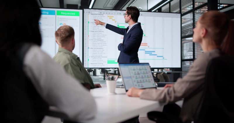A career coach and resume writer in business attire presents data on a large screen to three colleagues, one using a laptop, in a modern office meeting room.